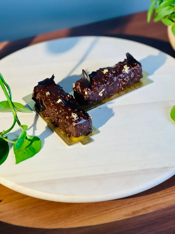 Two chocolate pastries with gold leaf on a pale wooden plate